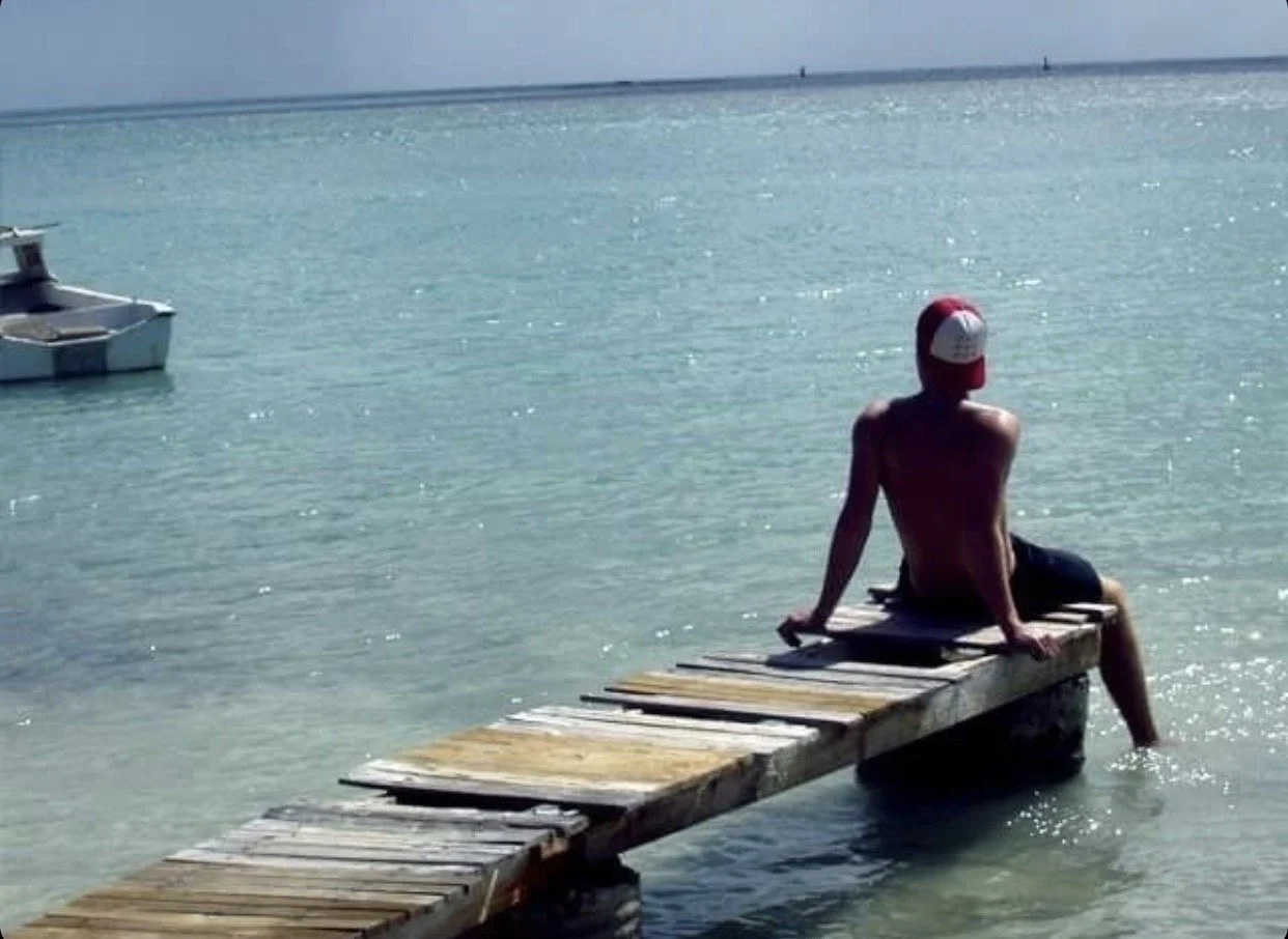 A person wearing a red and white cap is sitting on a wooden dock extending into clear blue water, with a boat nearby and a horizon in the background.