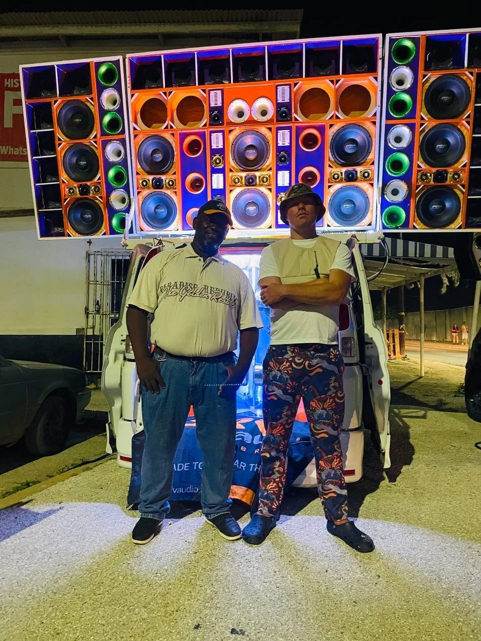 Two men standing in front of a large mobile sound system with multiple colorful speakers. The man on the left is wearing a white polo shirt, jeans, a hat, and sneakers. The man on the right is wearing a white t-shirt, patterned pants, and a hat. The 