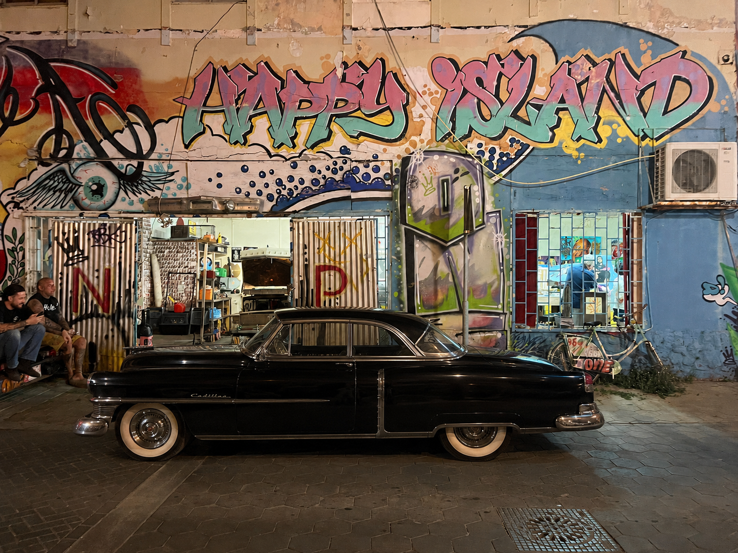 A vintage black Cadillac parked in front of a colorful graffiti mural on a building wall. The graffiti reads 'Happy Island' with vibrant and artistic designs, including a large eye with wings and abstract patterns. Two people are sitting on the left 
