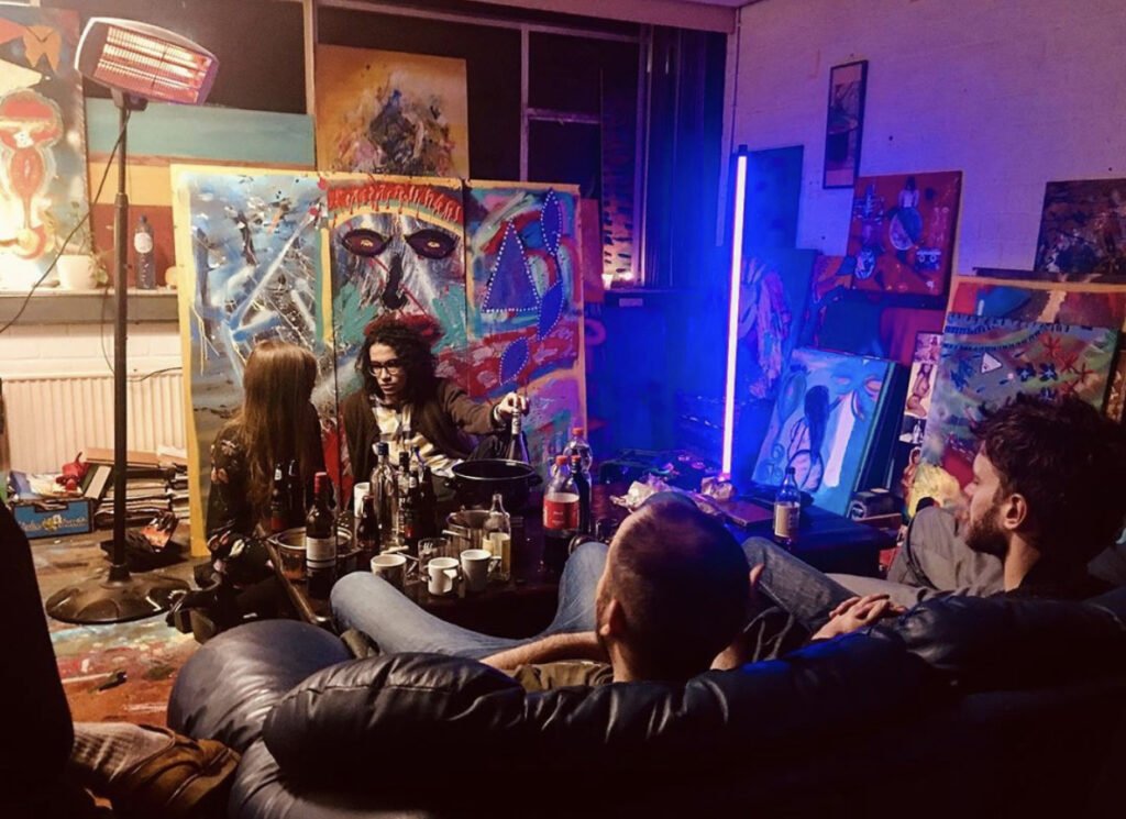 People sitting on a dark leather couch in an art studio or gallery, watching a woman with curly hair and glasses paint on an easel. The room is filled with colorful abstract artwork, bottles, and cups, with a bright blue vertical light and a red hue from a floor lamp.