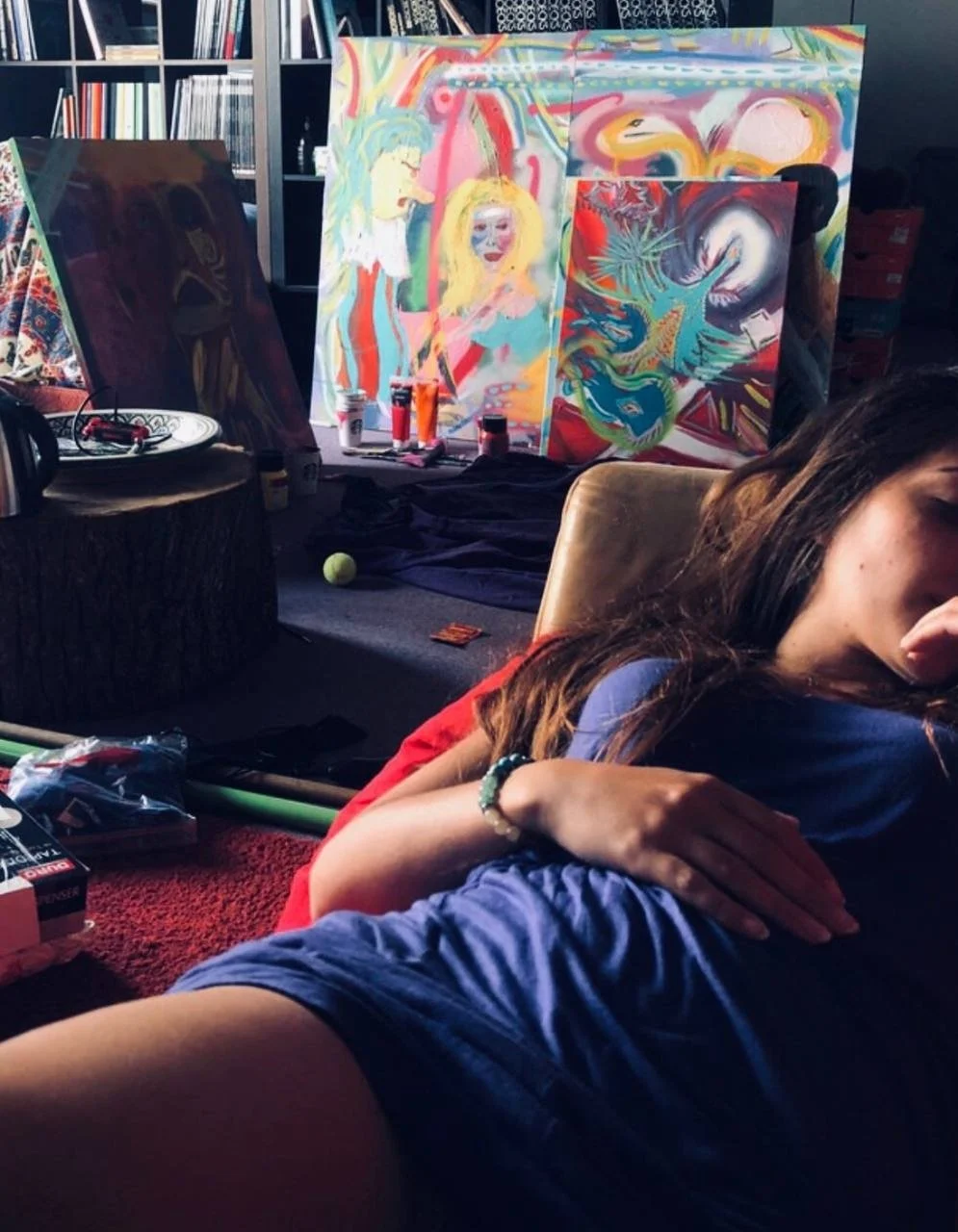 A young woman is lying down with her eyes closed in front of colorful abstract paintings and art supplies, including paint cans, a tennis ball, and brushes, in a cozy room with shelves of books.