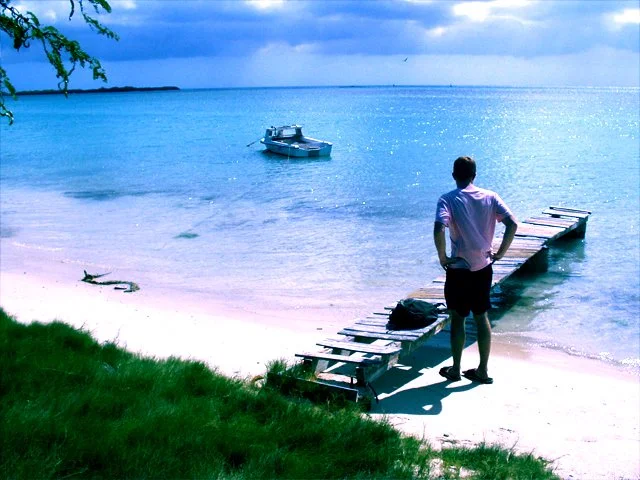 A man stands on a wooden dock by a calm, blue body of water with a boat floating nearby. There is a grassy area in the foreground and cloudy sky above.