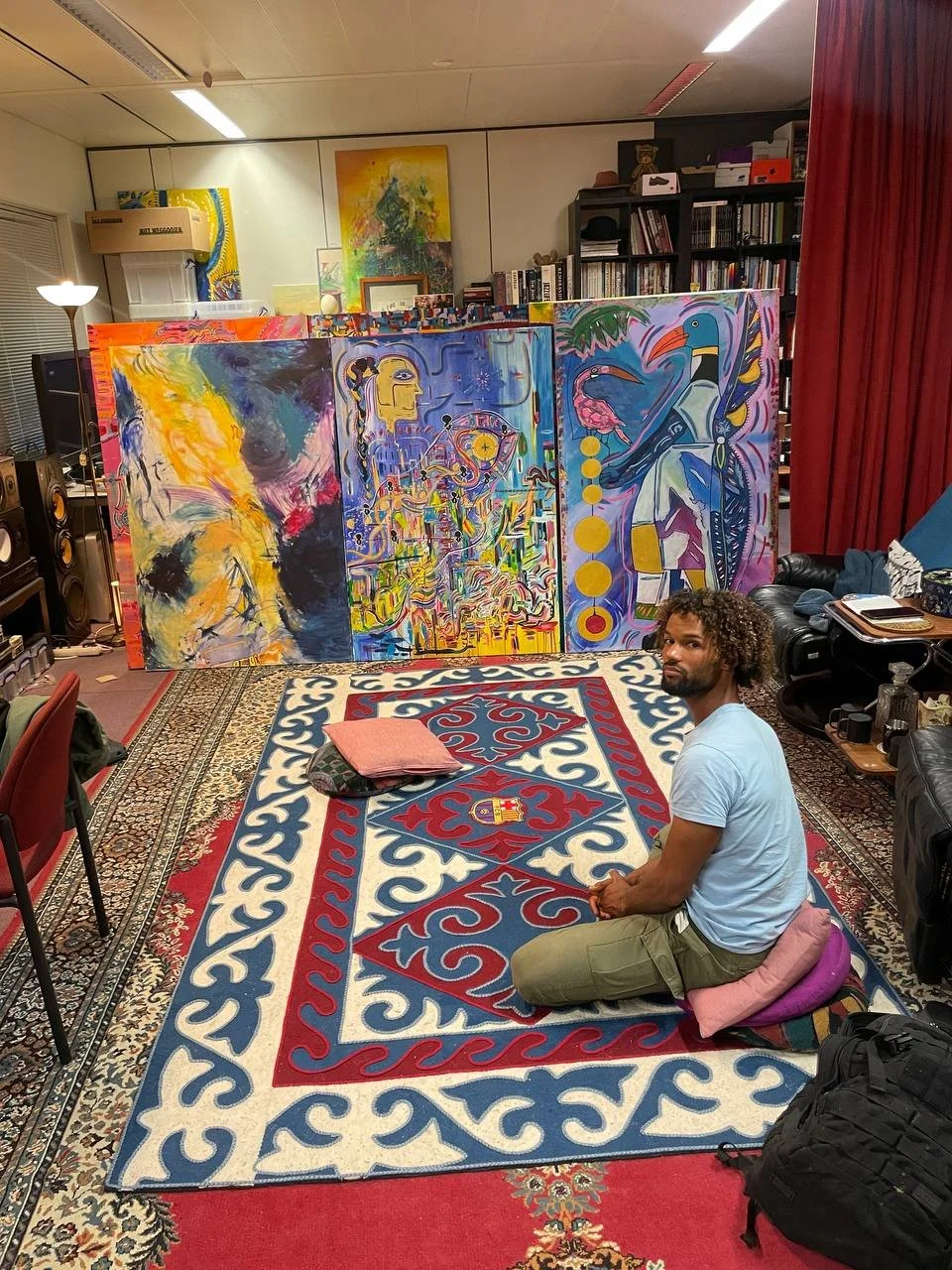 A man kneeling on a colorful Persian rug in a room with abstract paintings and bookshelves in the background.