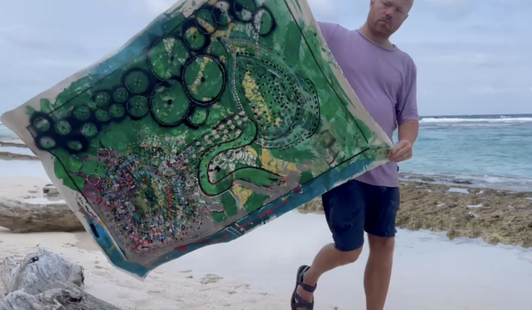 A man in purple shirt and shorts holding a colorful abstract painting on a beach with ocean waves in the background.
