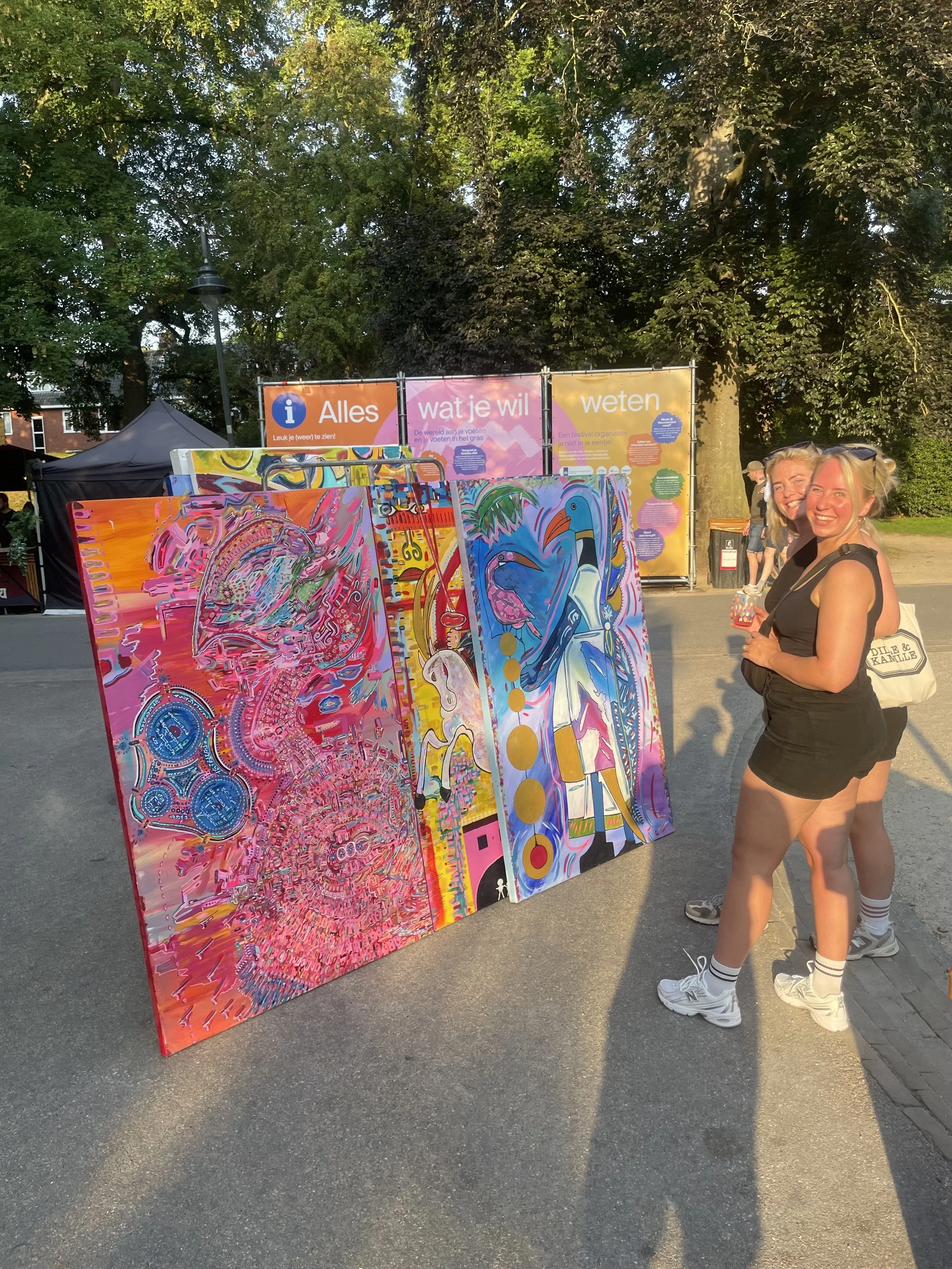 Two women smiling and posing near colorful abstract paintings at an outdoor art event in a park, with trees and informational banners in the background.