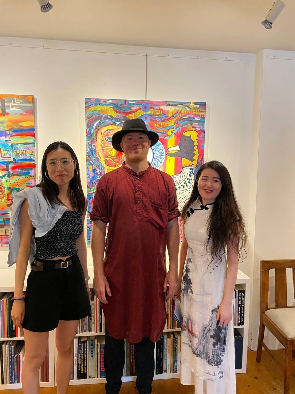 Roland Carter with fans from China standing in front of his colorful abstract paintings at a gallery exhibition, with bookshelves and a wooden chair nearby.