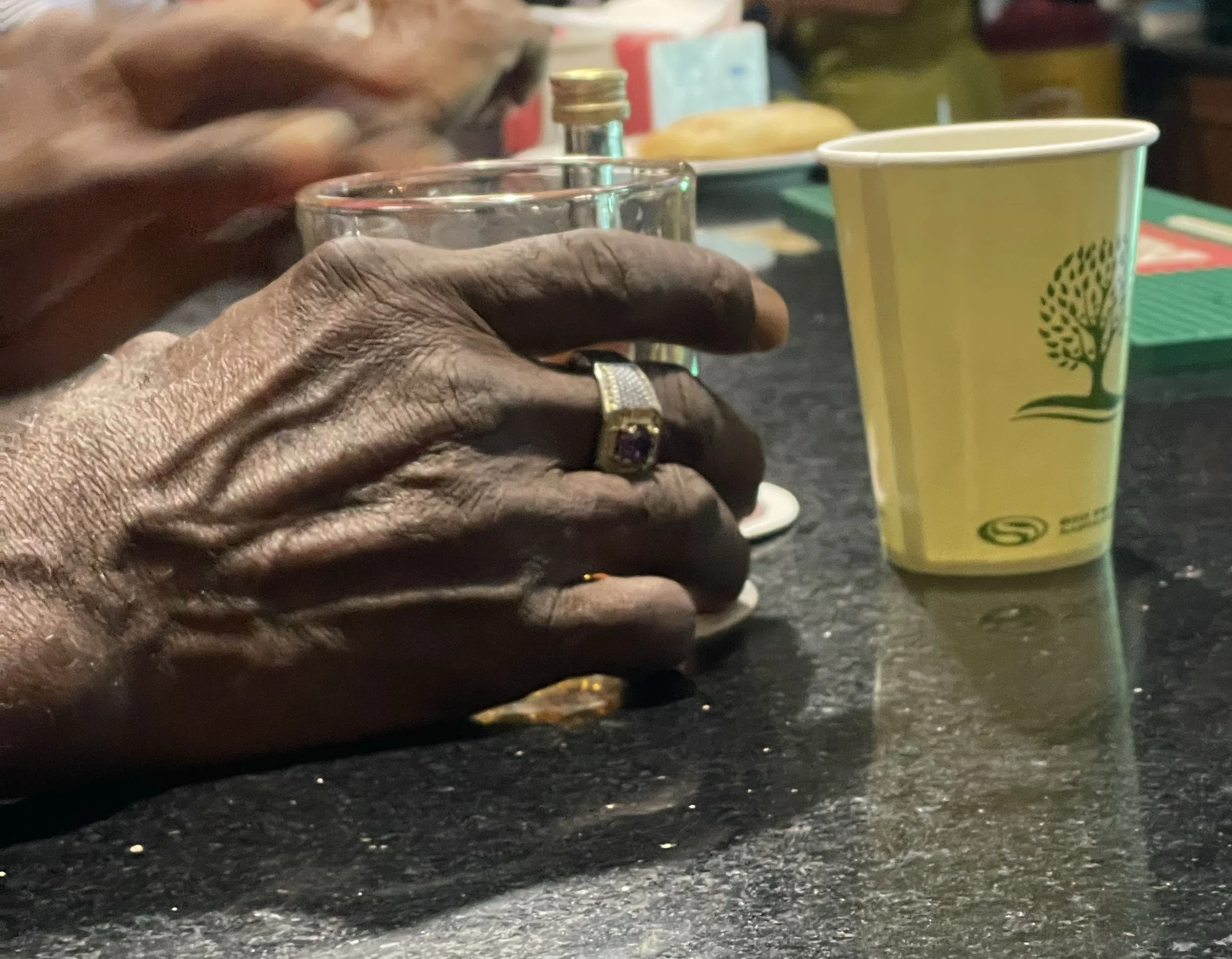 Close-up of a person's hand with a gold ring, resting on a black table. In the background, there is a yellow and green paper cup with a tree logo, a glass, a pepper shaker, and some packaging items.