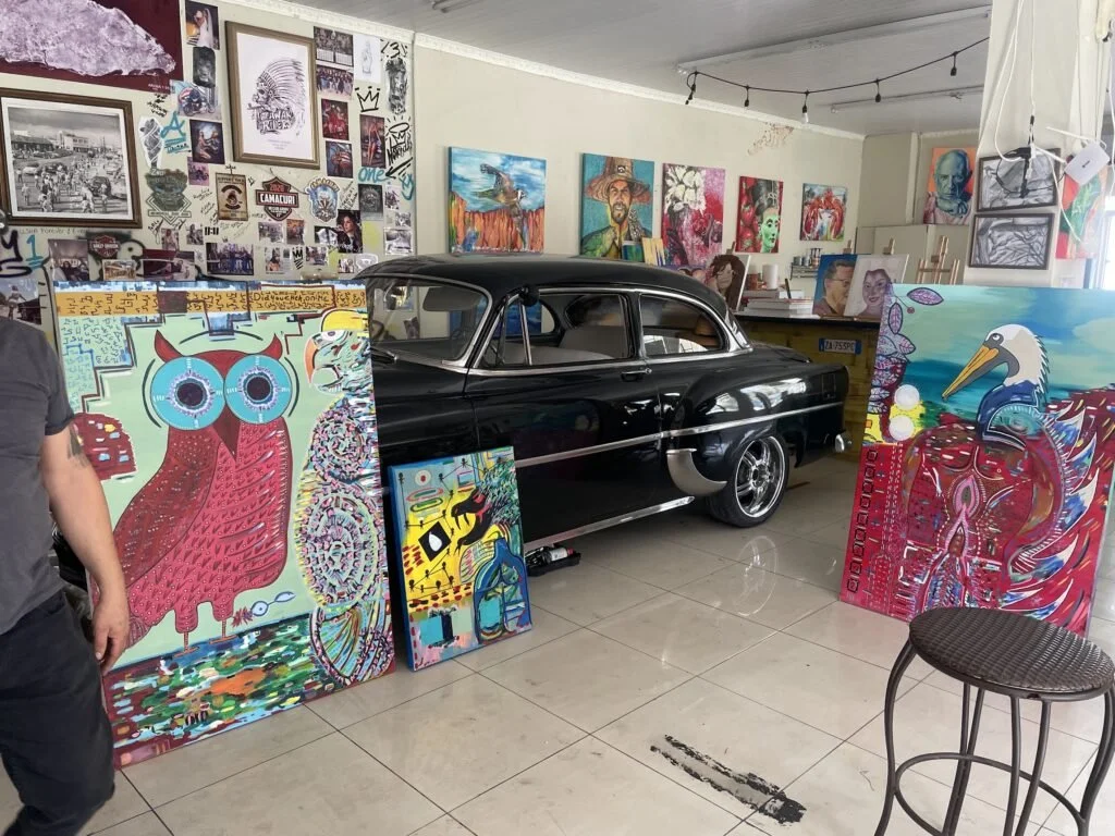 A collection of colorful psychedelic-style paintings, including an owl with large glasses, a toucan, and abstract patterns, displayed in an art studio with a vintage black car as a centerpiece.