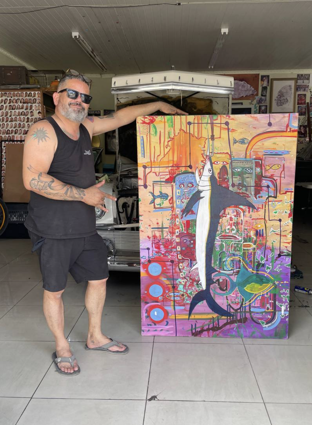 A man with tattoos, wearing sunglasses, a black sleeveless shirt, shorts, and flip-flops, is standing in front of a colorful abstract painting that features a whale and various cartoonish characters and patterns. The man is smiling, pointing at the a