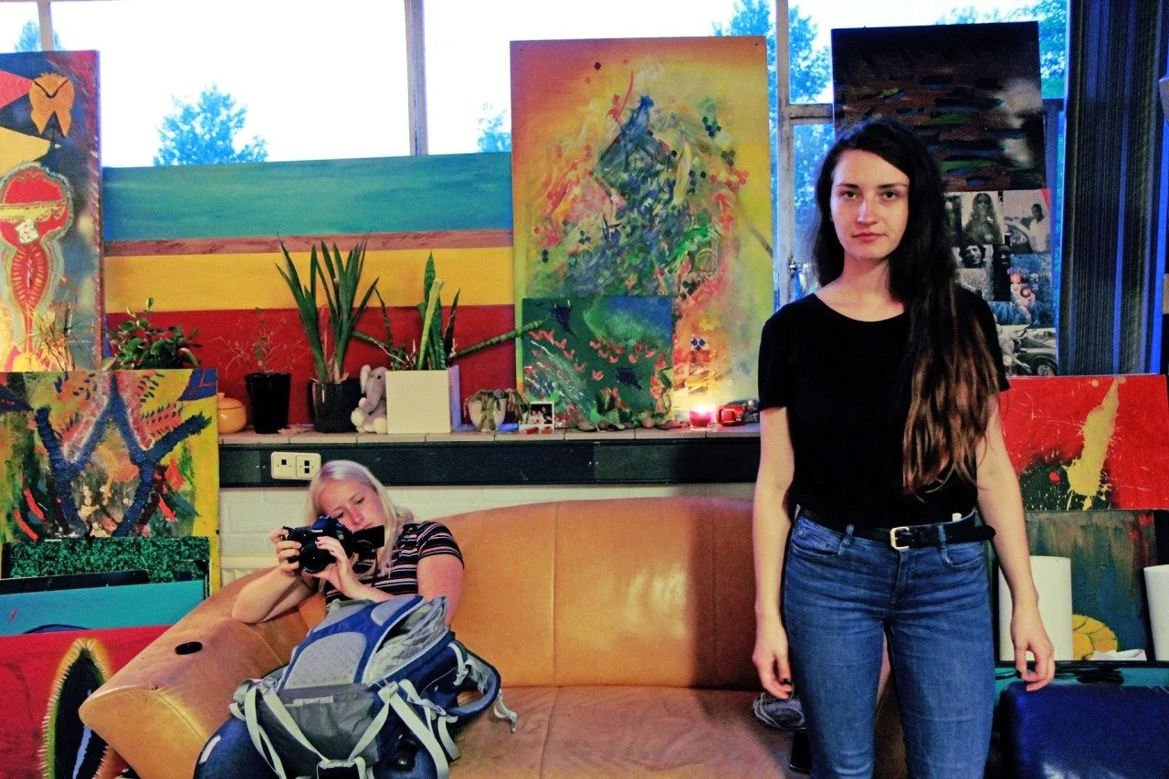 Two young women in an art-filled room. One woman with blonde hair is sitting on a tan couch, holding a camera, with a backpack in front of her. The other woman with long dark hair is standing next to the couch, wearing a black shirt and blue jeans. The background features colorful abstract paintings, potted plants, and a window with trees outside.