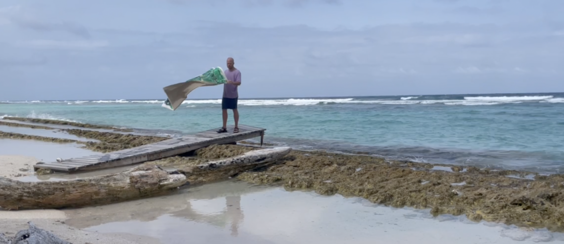 A person standing on a wooden dock near the rocky shoreline of a blue ocean, holding a green and beige object, with a cloudy sky above.