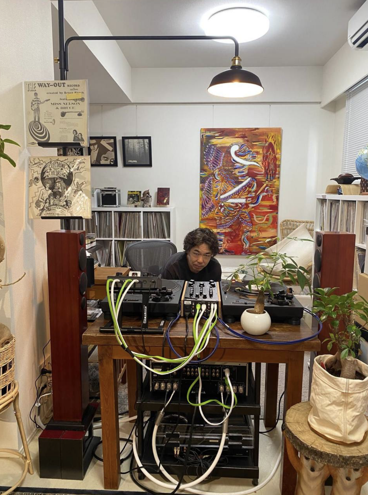 A man with dark curly hair sitting at a DJ setup with turntables and audio equipment in a cozy, decorated room with artwork and potted plants.