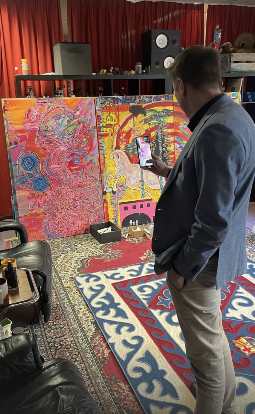 A man in a gray blazer and beige pants standing in a room with red curtains, colorful abstract artwork on a foldable partition, a patterned rug, black speakers, and various items on shelves and tables, taking a photo of the artwork with his smartphone.