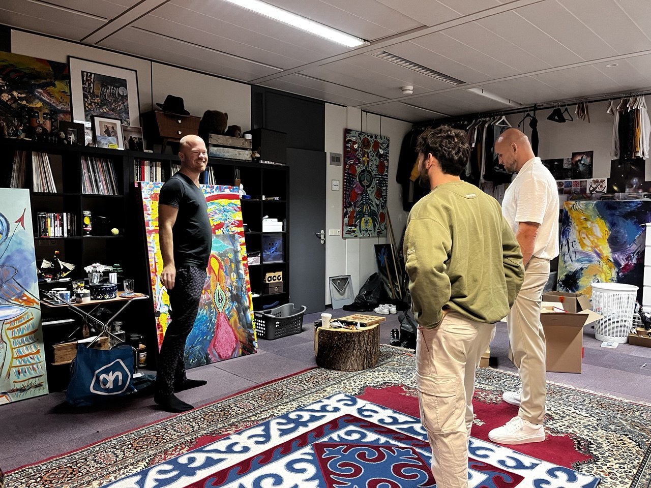 Three men in an art studio discussing colorful abstract paintings. One man is smiling, standing next to a large painted canvas, while the other two look on. The studio is decorated with shelves of books and art supplies, and wall art.