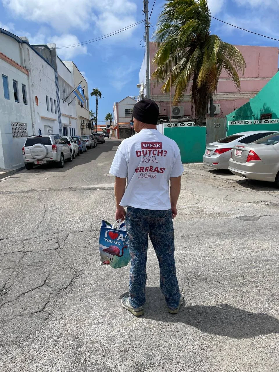 A man walking on a street with parked cars and buildings in the background, holding a grocery bag with a flamingo illustration, wearing a white T-shirt with text about speaking Dutch and a black beanie.