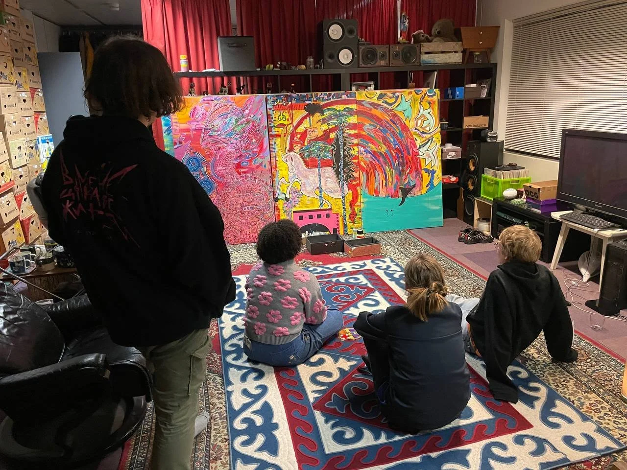 Four people in a room watching abstract paintings on a panel. One person is standing while three are seated on a patterned rug. The room has speakers, a TV, and colorful decorations.