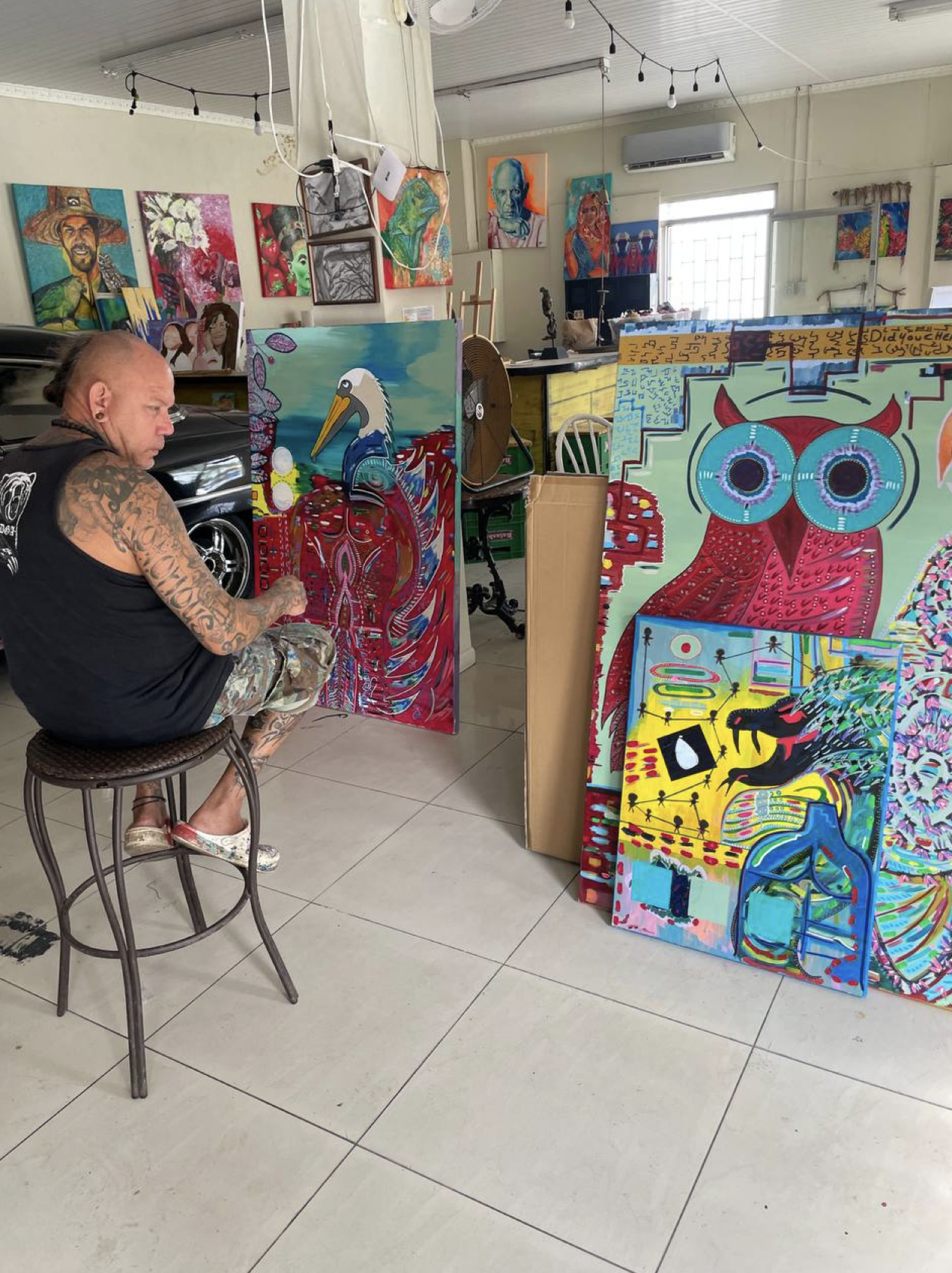 An artist sitting on a stool in an art studio surrounded by colorful paintings, including a drawing of a stork, abstract designs, and a creature with large eyes and red horns.