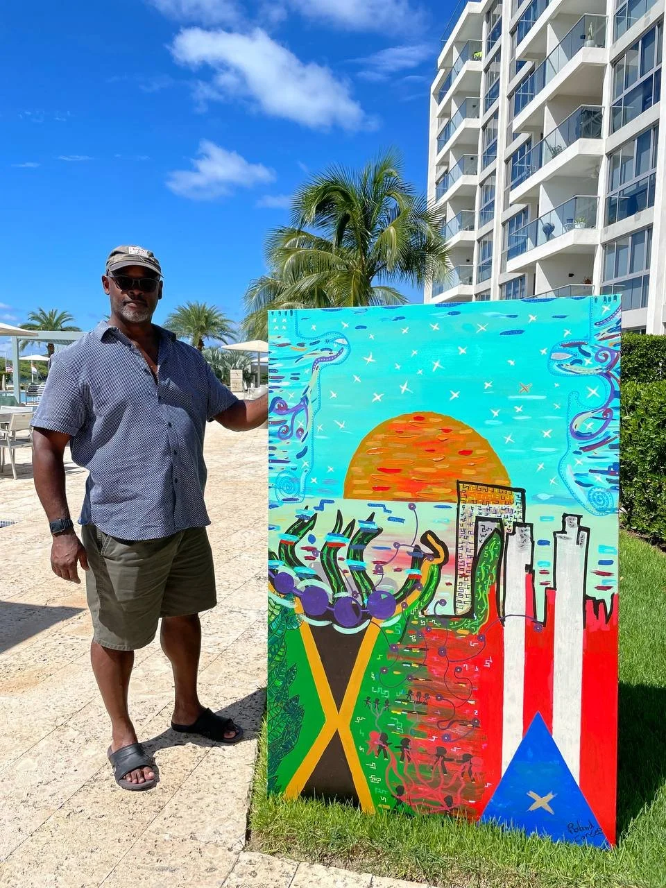 A man wearing sunglasses, a cap, and casual clothing standing next to a colorful street art painting outdoors near a beach or pool area with palm trees, a high-rise building, and a blue sky in the background.