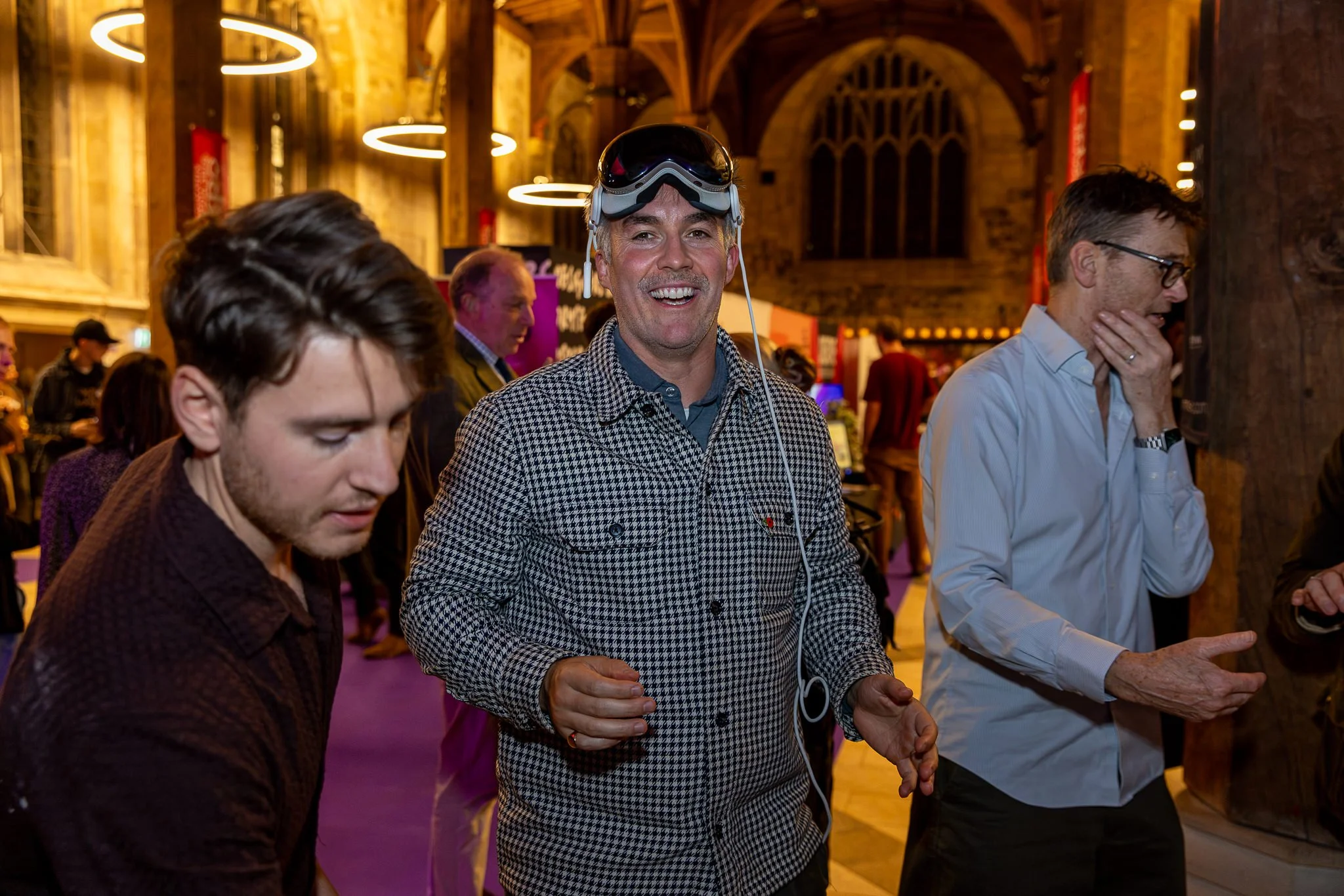 A group of people gathered in an indoor space with warm lighting. In the center, a man wearing a checkered shirt and black goggles on his head is smiling. To his right, another man in a blue shirt is touching his face, and to the left, a person with 