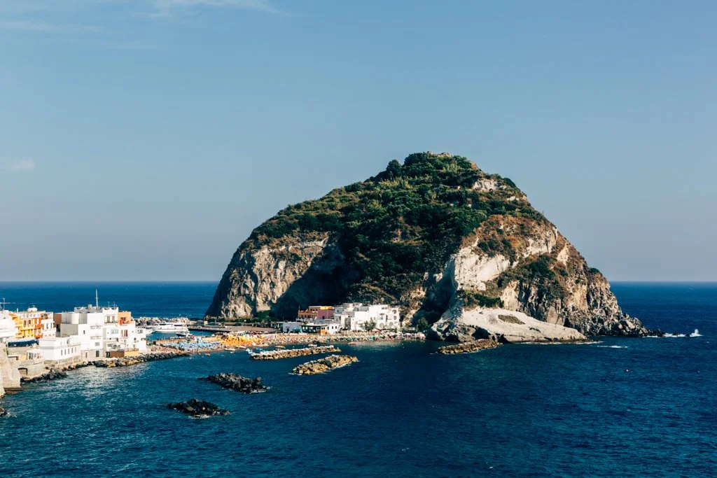 A large rocky island with green vegetation, situated next to a small coastal town with white buildings, and a body of water in the foreground.