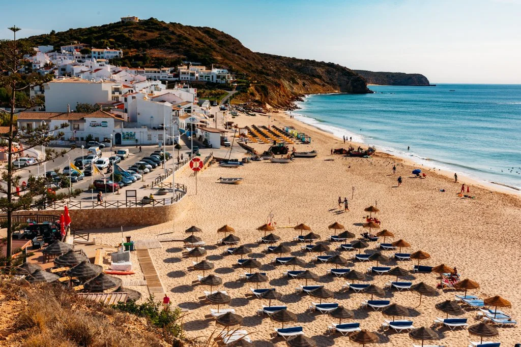 Sunny beach with rows of lounge chairs and umbrellas, people walking and relaxing, blue ocean water, white buildings on a hillside, and a small parking area near the shore.