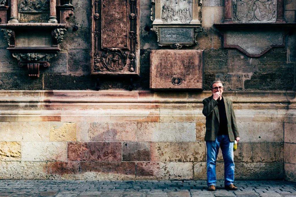 A man standing in front of a historic stone wall with several plaques and reliefs, holding a yellow object in his left hand, wearing glasses, a dark sweater, blazer, and jeans, with some fog or mist near the ground.
