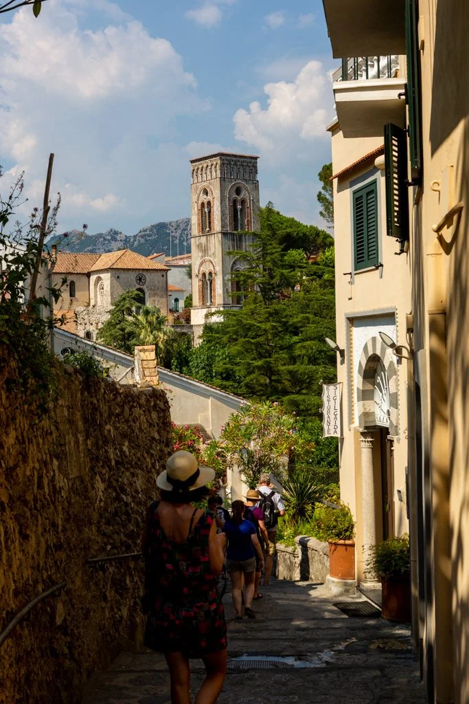 People walking down a narrow, inclined alleyway in a Mediterranean town, with a church tower and lush green trees in the background on a sunny day.