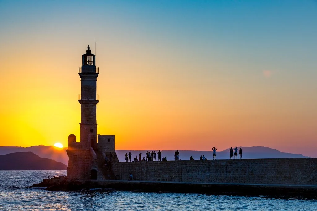 Lighthouse on a pier at sunset with people walking and taking pictures, distant mountains and water in the background.