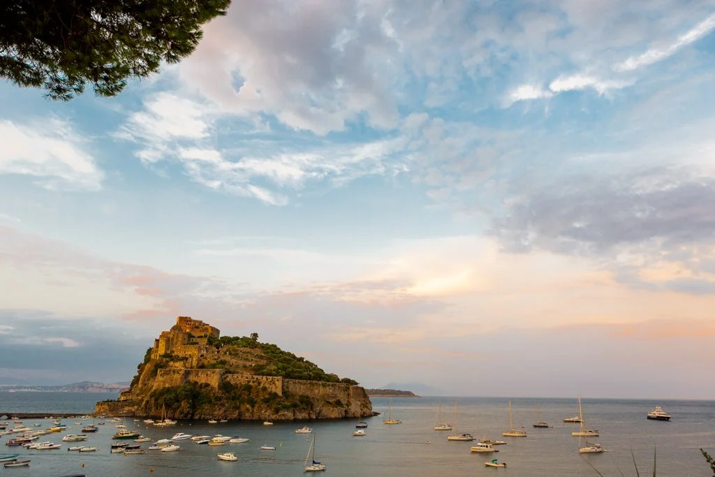 A castle or fortress on a hill surrounded by water with numerous sailboats and yachts docked, under a partly cloudy sky at sunset or sunrise.