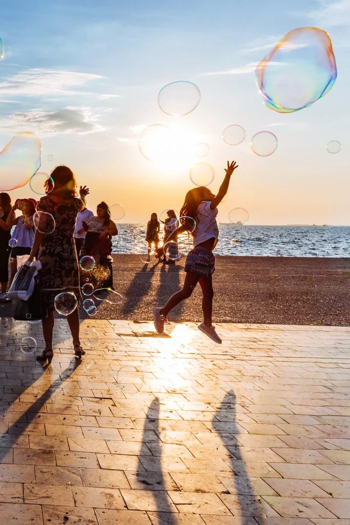 People at the beach during sunset, playing with floating soap bubbles. A girl in shorts and a shirt is jumping, with the sun behind her, casting long shadows on the paved walkway.