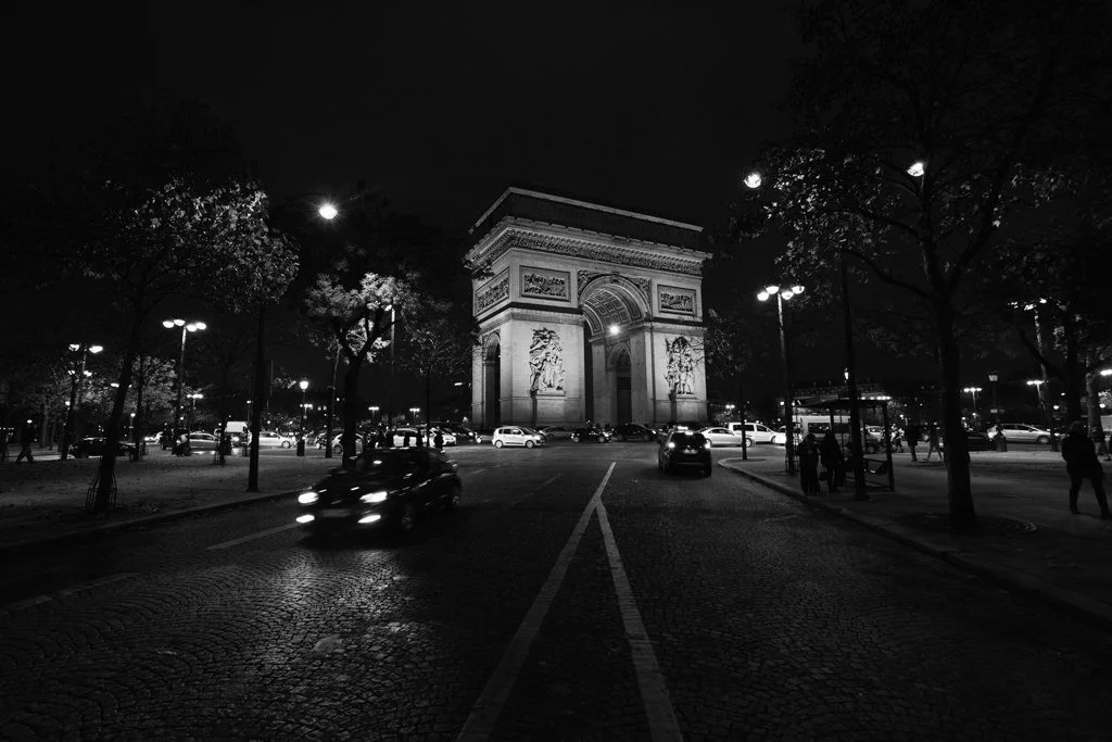 Nighttime view of the Arc de Triomphe in Paris with cars and pedestrians around, illuminated by streetlights.