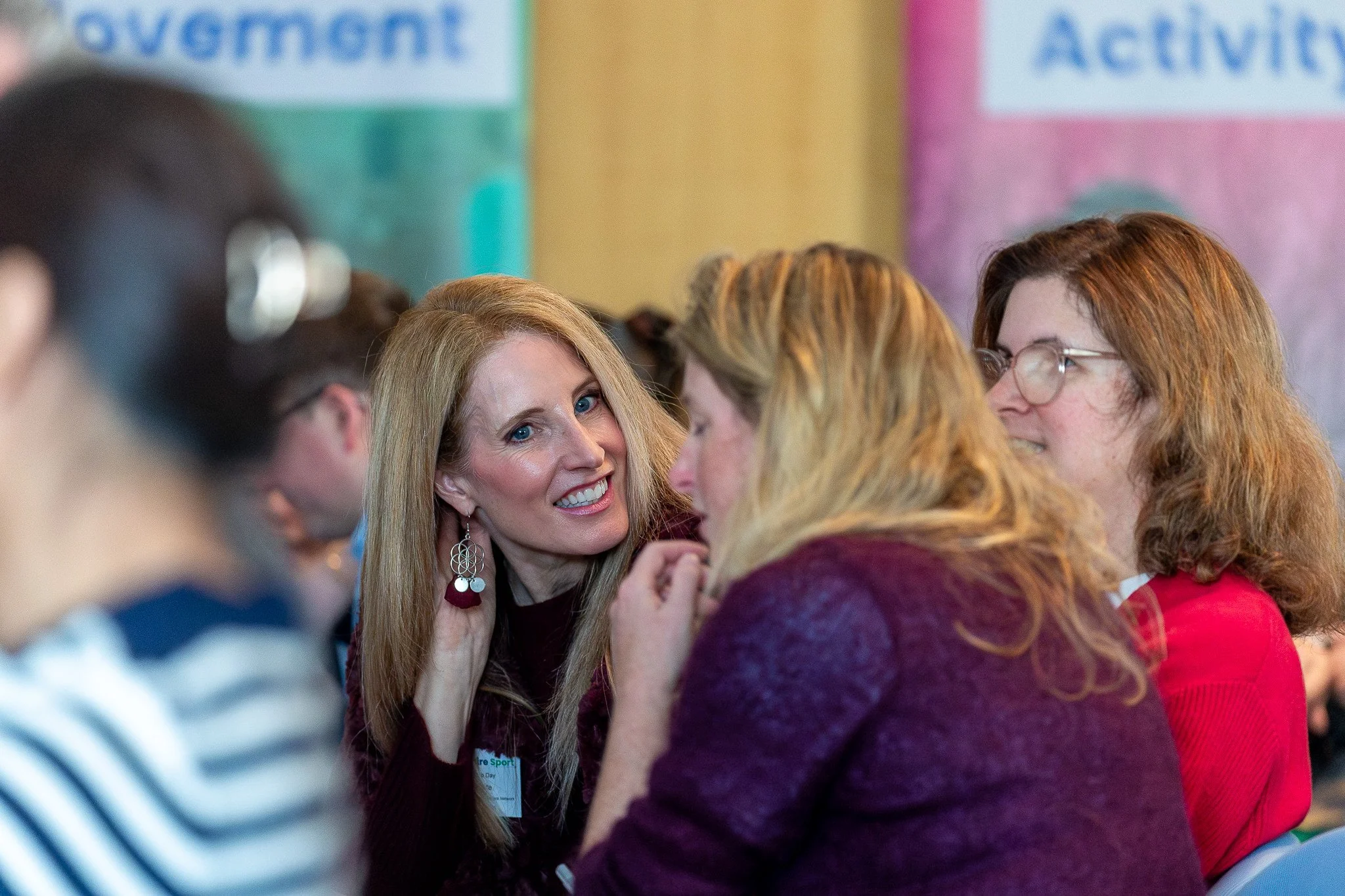 A group of people engaged in conversation, with a woman in the center smiling and wearing earrings and a name badge. Others around her are talking and listening, with signs in the background indicating a professional or conference setting.