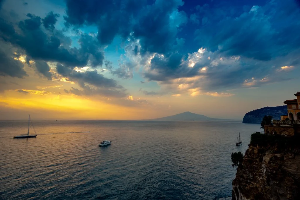 Sunset over the ocean with boats, clouds, and a distant mountain, with buildings on the rocky coastline on the right.