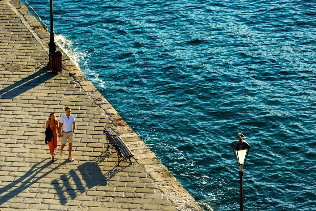 A couple walking along a stone pier by the water, with a lamppost and benches, cast shadows on the ground.