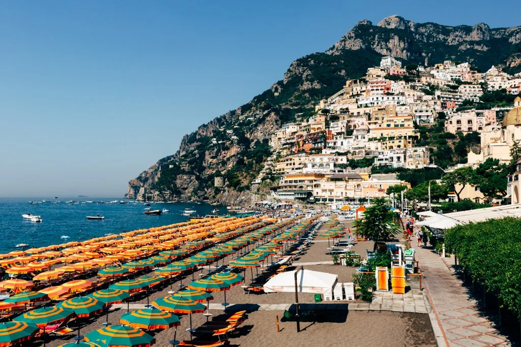 Beach with colorful umbrellas and lounge chairs, boats on the water, hillside town with white buildings, and a pathway with trees and people.