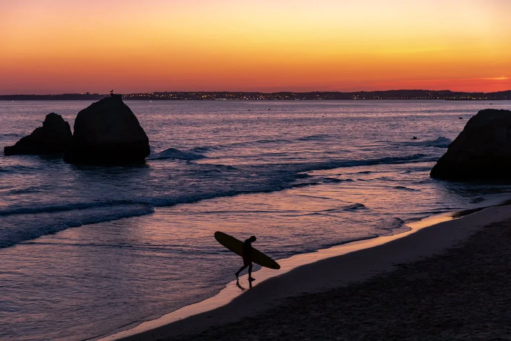 A person carrying a surfboard walking along a beach at sunset with large rocks in the water and a city skyline in the distance.
