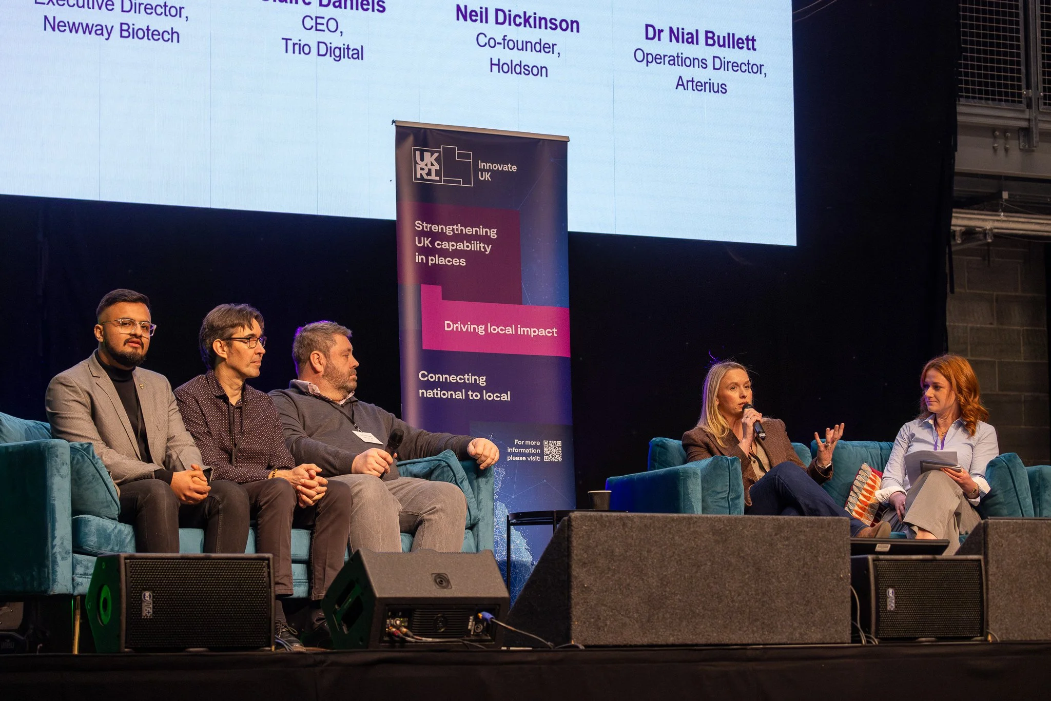 A panel discussion with five people sitting on a stage, women and men, some holding microphones and notes. Behind them is a large screen and a banner displaying the UKRI logo and messages about strengthening UK capabilities and driving local impact.