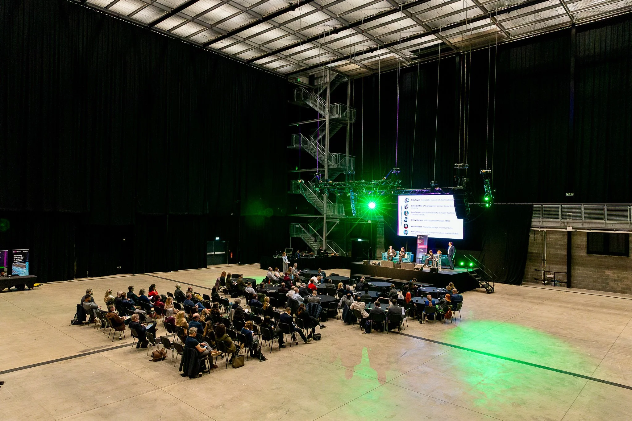 A large indoor conference or event space with a stage and audience seating. The stage has a large screen displaying presenters' names and titles, and a panel of speakers seated. The room features high ceilings, dark curtains, and stage lighting, with