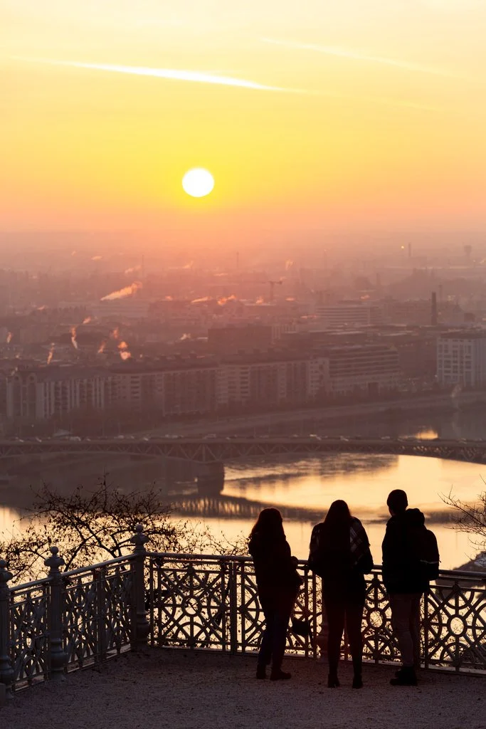Three people standing on a balcony overlooking a city river at sunset.