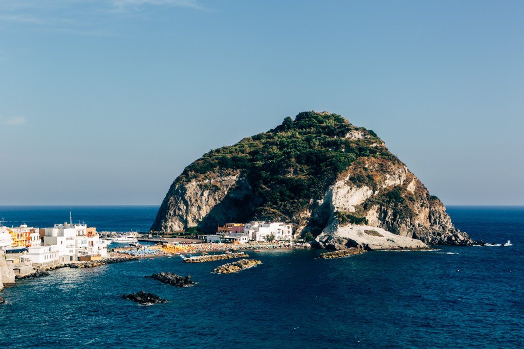 A large rocky island with green vegetation on top, located off a coast with white buildings, blue water, and a clear sky.