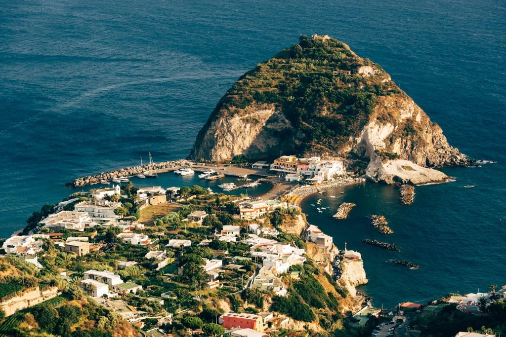 A coastal hilltop village with white buildings, a marina with boats, and a large rocky hill covered in greenery overlooking the ocean.