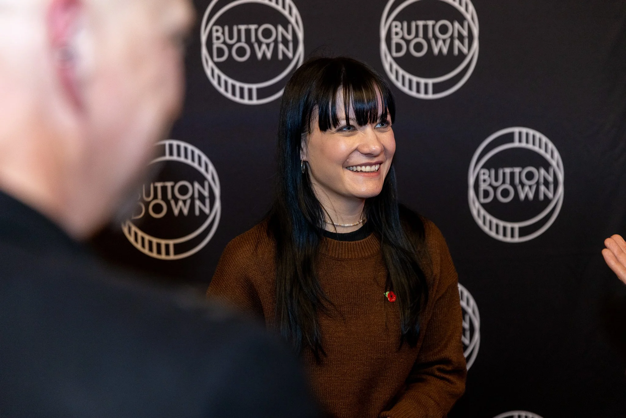 A woman with black hair and bangs, smiling and wearing a brown sweater, standing in front of a black background with white Circular logos that say 'Button Down'.
