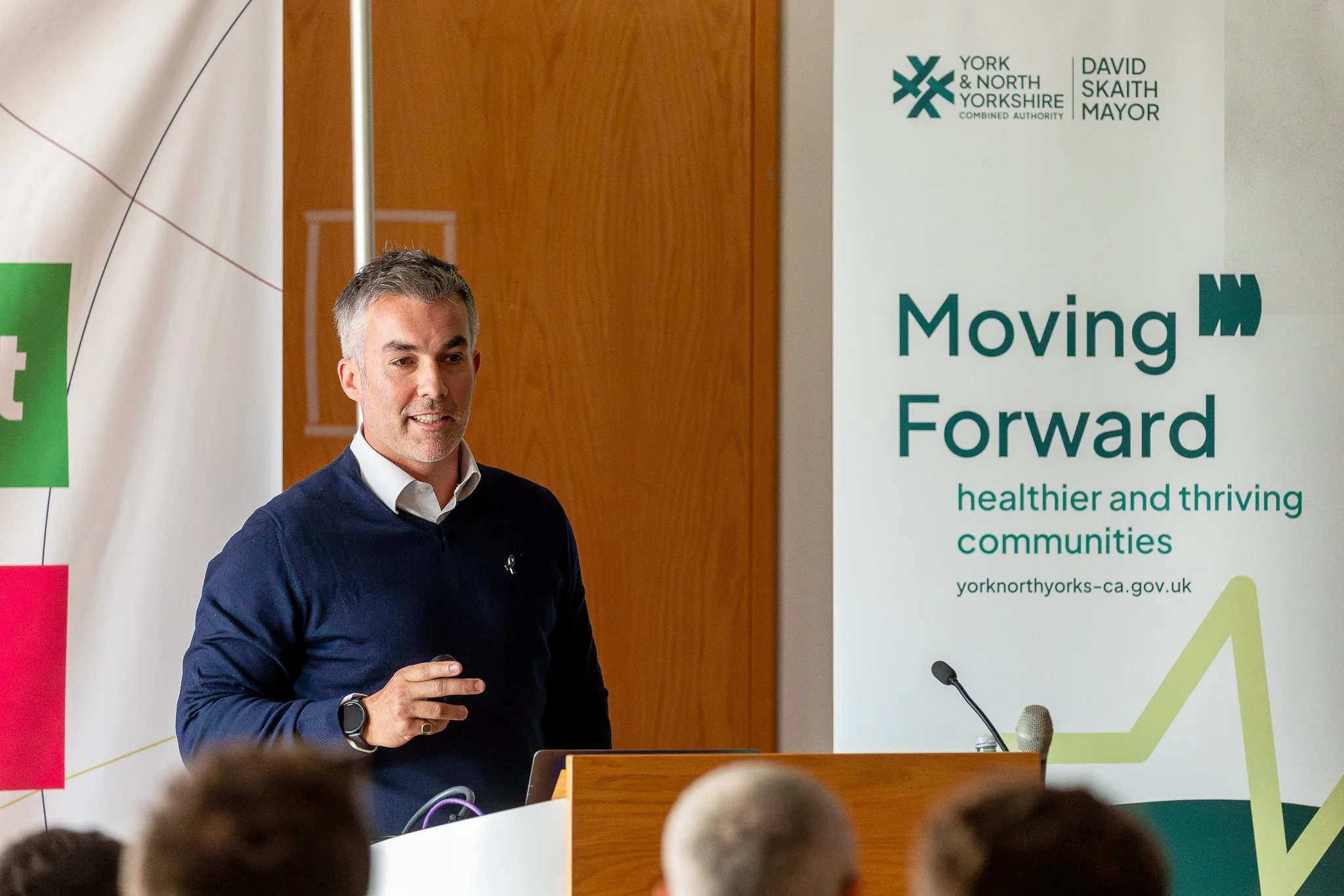 A man with gray hair and a beard, wearing a navy blue sweater and a white shirt, speaking at a podium during a presentation or event. Behind him, there are banners and a wooden wall. One of the banners reads 'Moving Forward, healthier and thriving co