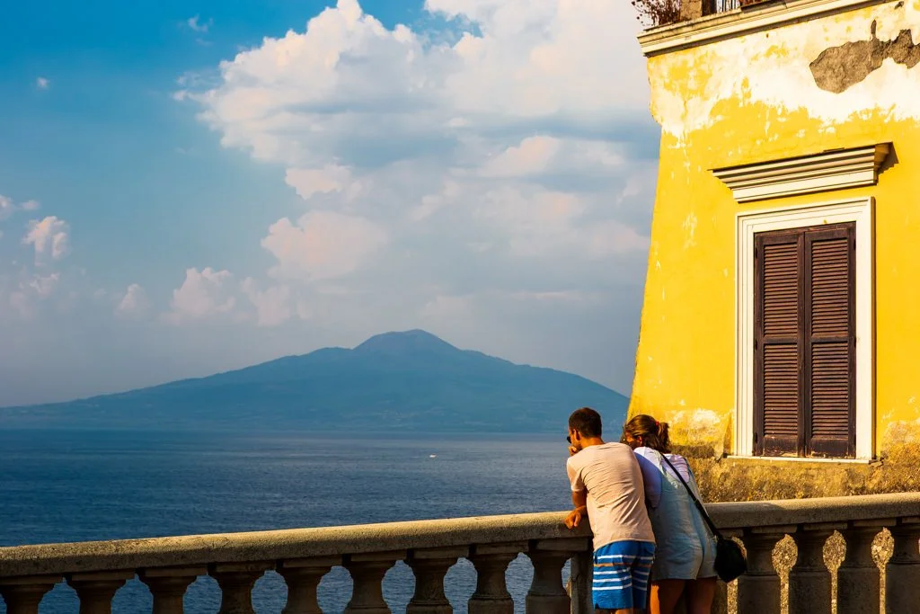 A man and woman leaning on a balcony, looking out over the ocean with an island or mountain in the distance, next to a yellow building with a closed window shutter.