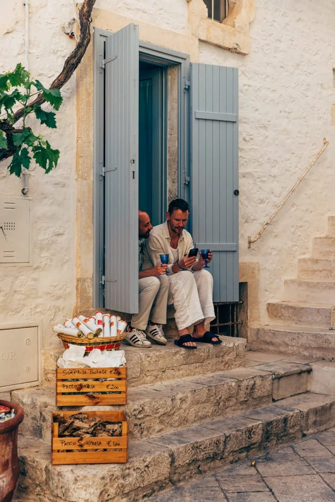 Two men sitting on stone steps in front of a house with light blue shutters, looking at a phone and drinking from blue cups, with a basket of paper tubes and wooden boxes with handwritten signs for surprise packages outside.