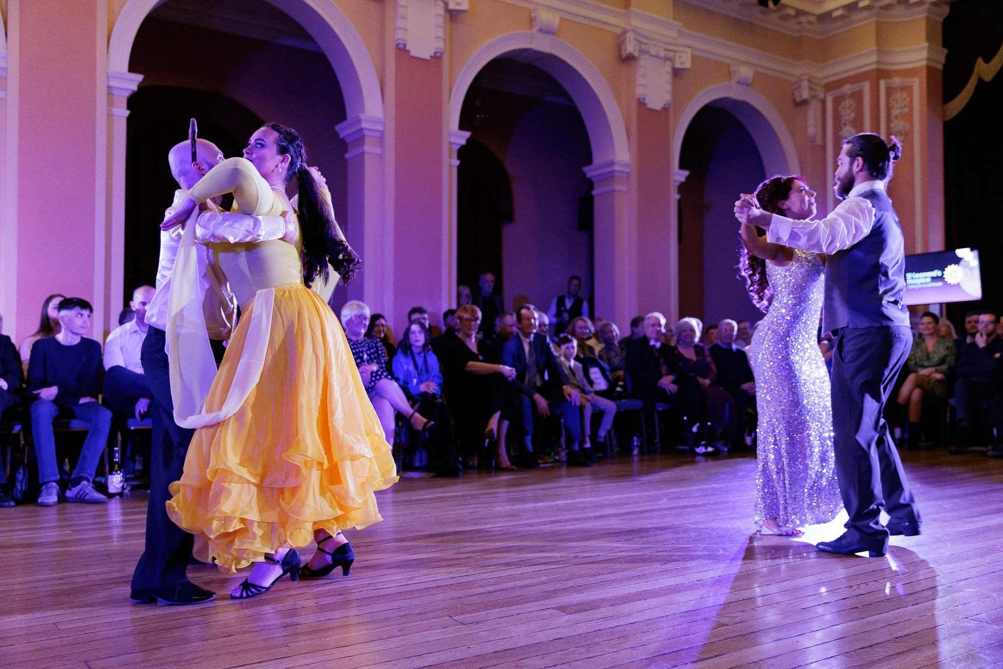 Two couples dancing ballroom in a grand, ornate room with pink walls and high arches. The women are wearing elegant evening gowns, and the men are in formal attire. Audience seated around the dance floor watching the performance.