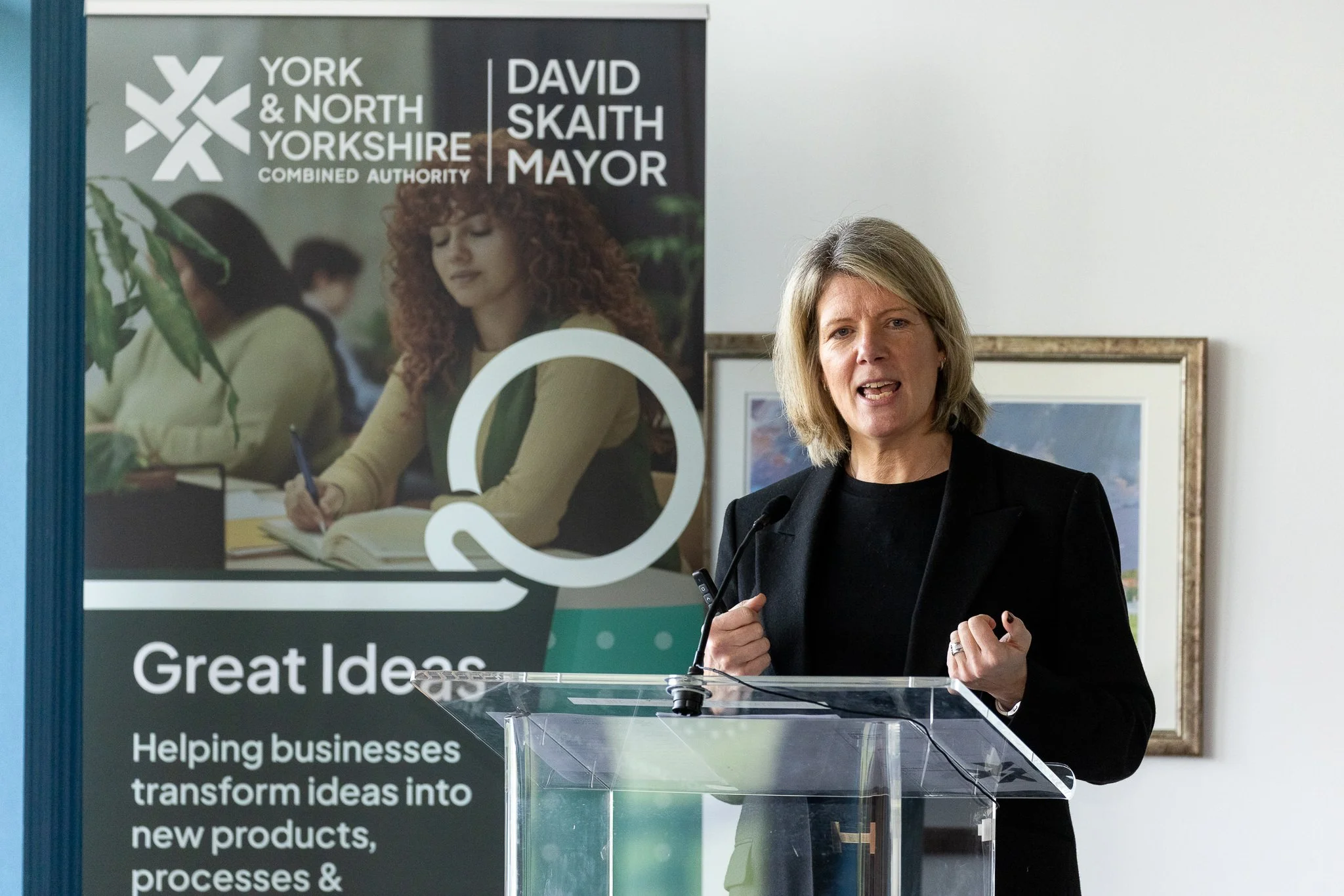 A woman speaking at a podium with a microphone in front of a banner that reads "York & North Yorkshire Combined Authority" and "David Skaith Mayor". The woman is wearing a black blazer and has shoulder-length blonde hair.