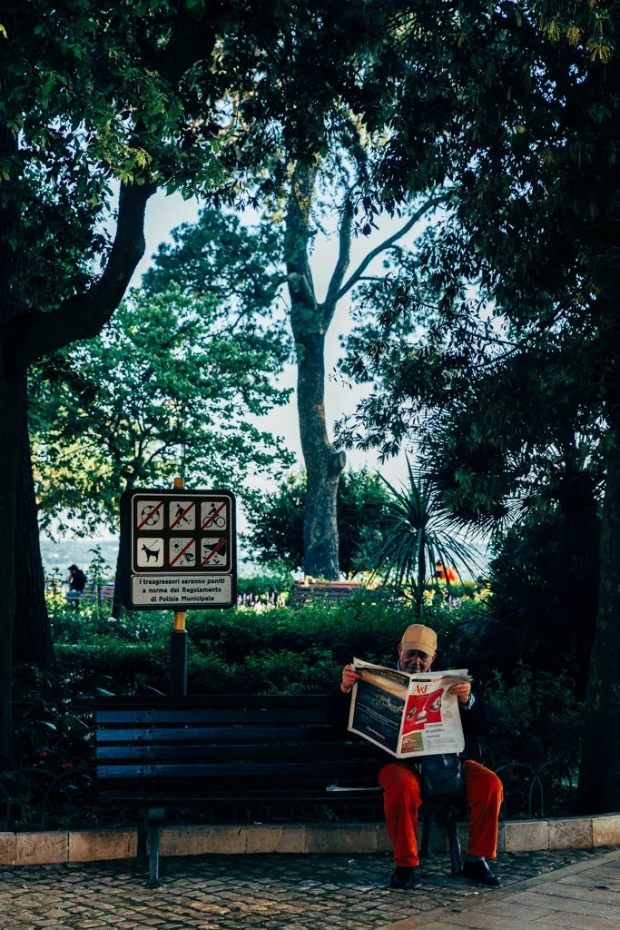 A man wearing a beige cap and red pants is sitting on a park bench while reading a newspaper. Behind him is a sign indicating no dogs, bicycles, or smoking allowed.