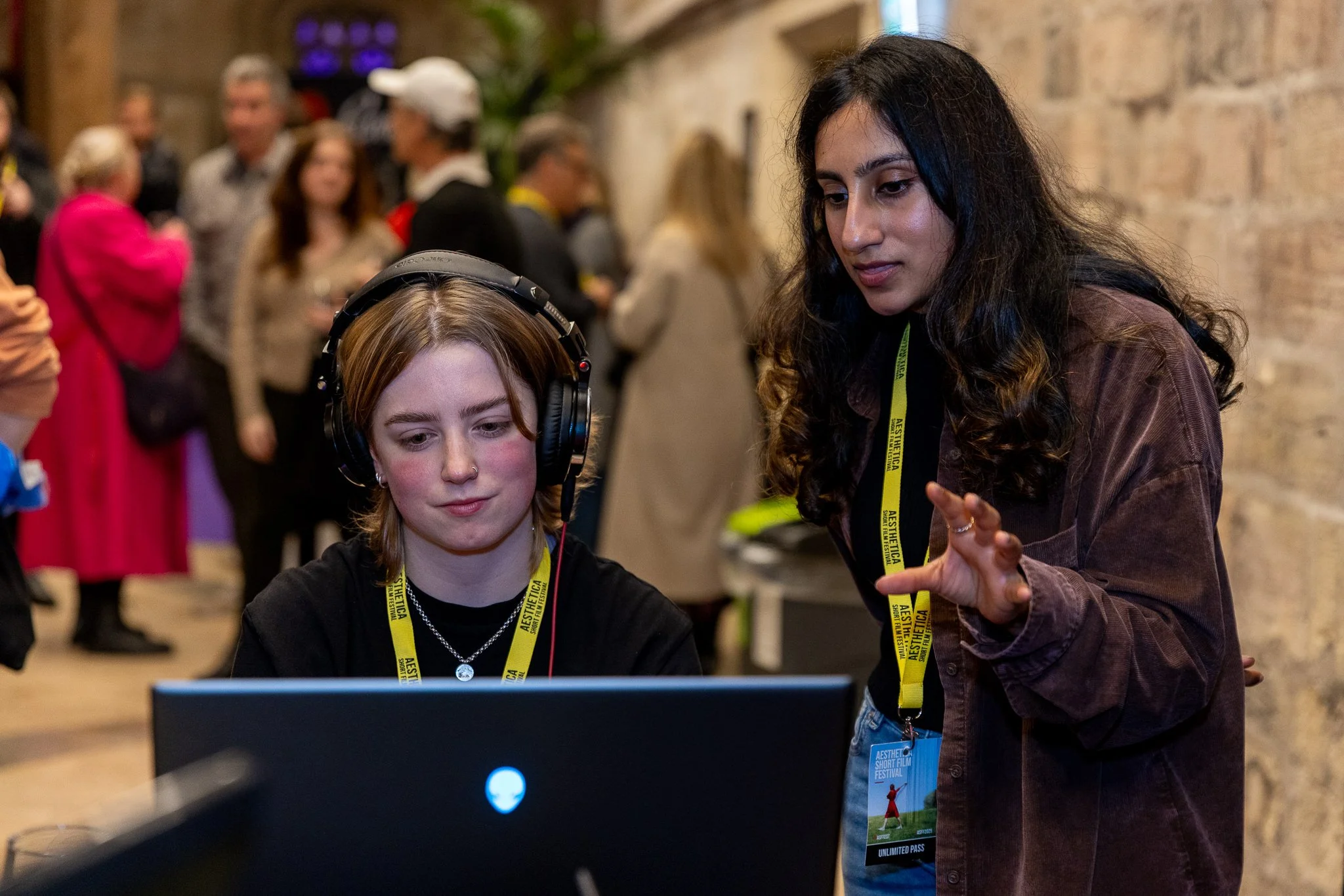 A young woman wearing large headphones and a black shirt is sitting at a computer, while an older woman with long dark hair in a brown jacket and an accreditation badge is explaining something to her. Both are wearing yellow lanyards with event info,