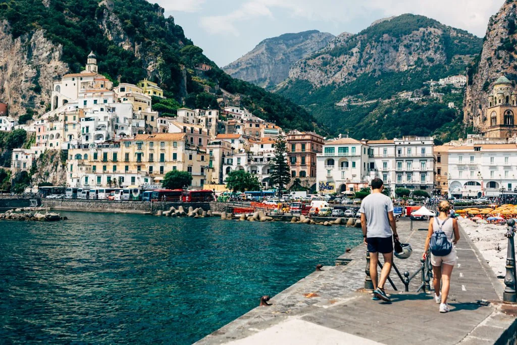 Couple walking on waterfront promenade with colorful hillside town and mountains in background